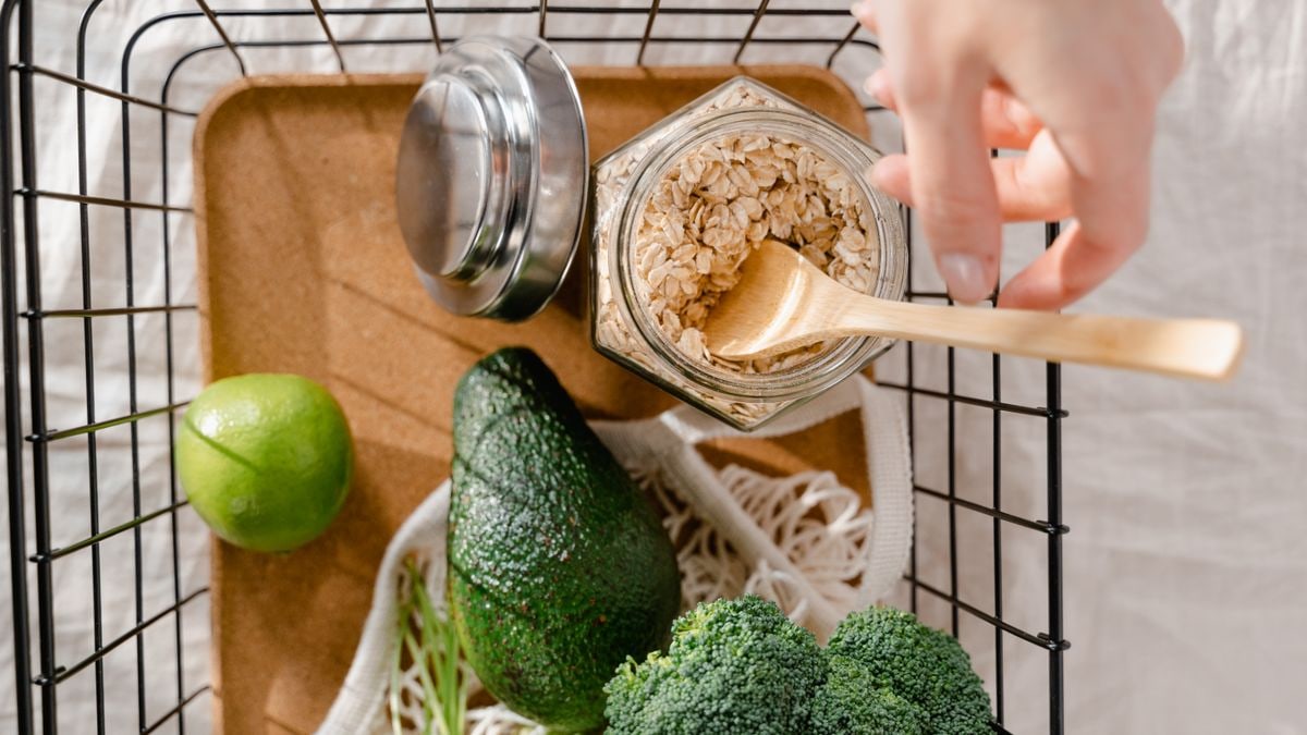 A metal basket with fresh vegetables, avocado, lime, and oats arranged in a flat lay style.